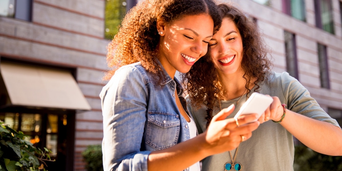Two smiling women stand outdoors in front of a building, looking at a smartphone together. They appear excited and engaged, as if browsing online for a home or shopping for a property.